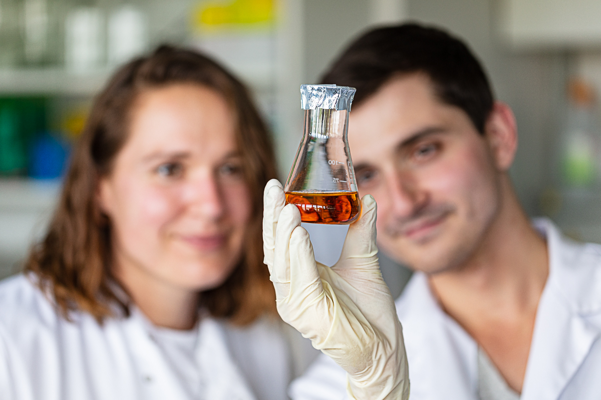 Two laboratory staff look at an Erlenmeyer flask with orange-reddish growth medium in the foreground.
